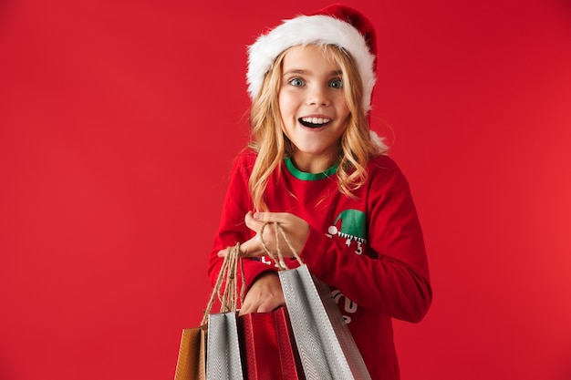 Joyeuse petite fille portant un chapeau de Noël debout isolé, portant des sacs à provisions