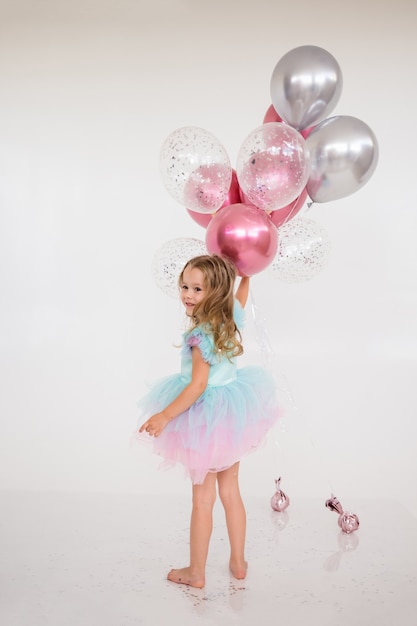 Joyeuse petite fille vêtue d'une robe de fête avec une jupe tutu tient un tas de ballons sur fond blanc avec une copie de l'espace