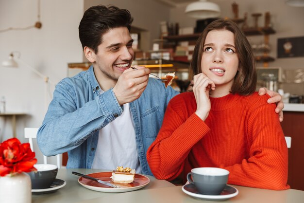 Joyeuse photo de jeune couple de 20 ans assis dans un café et homme nourrir la femme avec un gâteau savoureux