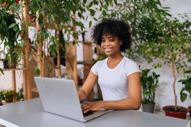 Joyeuse pigiste afro-américaine travaillant sur un ordinateur portable assis au bureau depuis une salle de bureau à distance avec un design moderne de biophilie