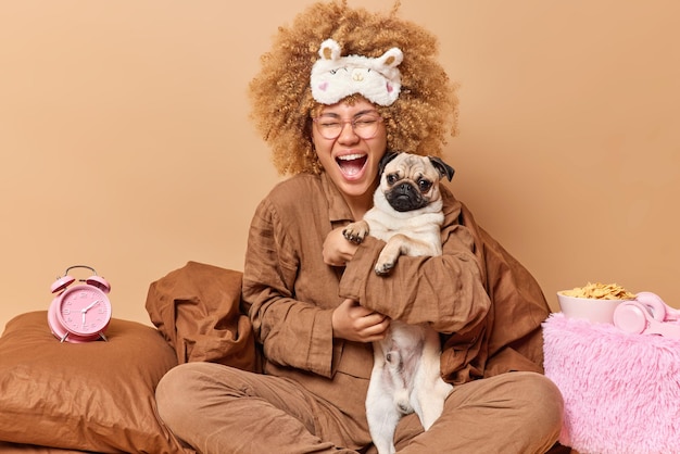 Joyeuse propriétaire d'un animal de compagnie pose dans un lit confortable avec un jeu de chien carlin et se prépare à dormir pose dans la chambre avec une couverture d'oreiller de réveil et un bol de cornflakes autour du concept d'animaux domestiques