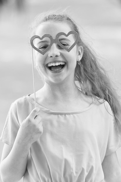 Photo joyeuse saint valentin petit enfant avec un sourire heureux et un regard drôle à travers des lunettes en forme de coeur bonne petite fille souriante avec des accessoires de photomaton sur un bâton célébrant la bonne journée internationale des enfants