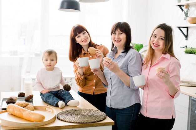 Joyeuse souriante séduisante jeunes femmes, femme d'âge moyen et petite mignonne petite fille mangeant des cookies et des cupcakes et buvant du café à la cuisine à domicile Concept de fête des mères heureux, cuisiner ensemble