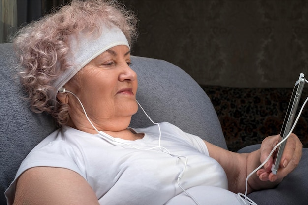 Photo joyeuse vieille femme écoutant de la musique via des écouteurs tout en étant assise sur une chaise après une séance d'entraînement.