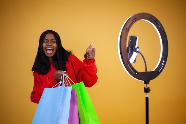 Une joyeuse vlogger afro-américaine dans un sweat à capuche rouge avec des sacs colorés, une lampe annulaire et un téléphone