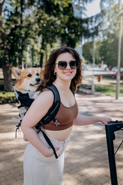 Une joyeuse voyageuse souriante chevauche son scooter électrique dans un parc de la ville avec un chien Welsh Corgi Pembroke dans un sac à dos spécial