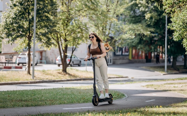 Une joyeuse voyageuse souriante chevauche son scooter électrique dans un parc de la ville avec un chien Welsh Corgi Pembroke dans un sac à dos spécial