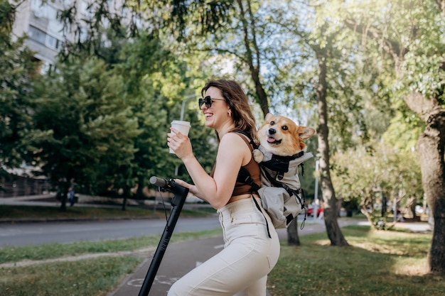 Une joyeuse voyageuse souriante se tient debout avec un scooter électrique et boit du café dans un parc de la ville avec un chien Welsh Corgi Pembroke dans un sac à dos spécial