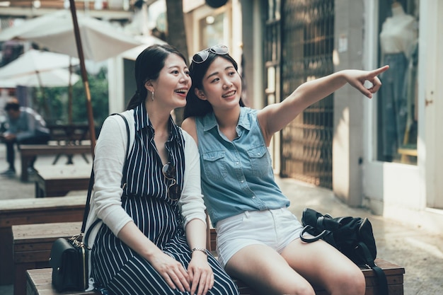 De joyeuses amies asiatiques bavardent alors qu'elles sont assises sur un banc dans la zone commerciale en plein air. la jeune fille pointe le doigt de côté et rit en montrant sa soeur. deux belles femmes partageant la vue sur la ville