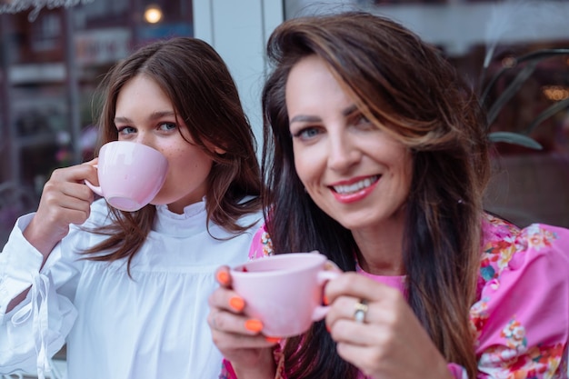 Joyeuses belles dames assises ensemble à l'extérieur dans un café et appréciant de boire du thé dans des tasses roses en gros plan fête de vacances célébration événement amitié meilleures amies femmes séduisantes