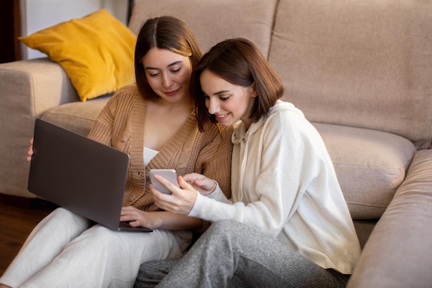 De joyeuses dames européennes du millénaire s'assoient sur le sol pour regarder une vidéo sur un ordinateur portable et un smartphone dans un salon confortable