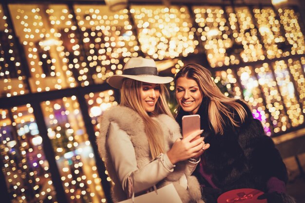 Joyeuses deux sœurs s'amusant dans la rue de la ville la nuit de Noël. Ils utilisent un smartphone et parlent de quelque chose de drôle.