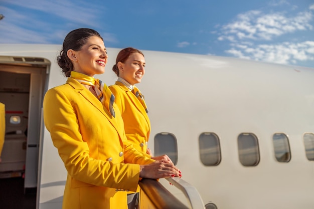 Joyeuses hôtesses de l'air en uniforme d'aviation regardant ailleurs et souriant tout en se tenant près de la porte de l'avion