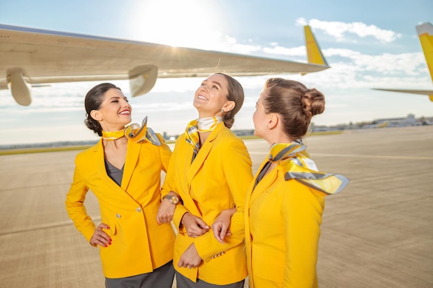 Joyeuses hôtesses de l'air en vestes jaunes regardant un collègue et souriant tout en se tenant à l'extérieur sur l'aérodrome