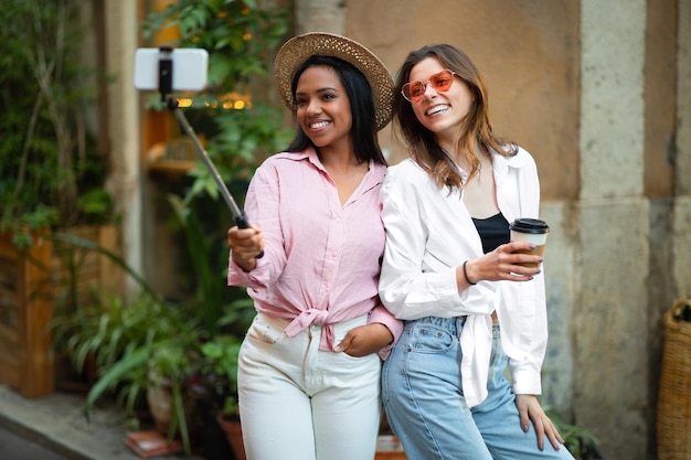 De joyeuses jeunes femmes multiraciales en tenue décontractée avec une tasse de boisson profitent d'un voyage dans la nouvelle ville prennent un selfie sur un smartphone