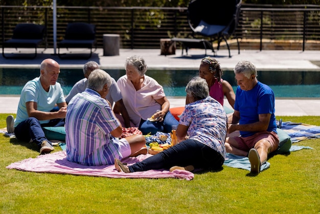 Joyeuses personnes âgées diverses assises sur une couverture et pique-niquant dans le jardin