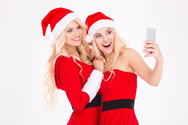 Joyeuses sœurs blondes jumelles en vêtements et chapeaux rouges du père Noël se cachant posant près de l'arbre de Noël sur fond blanc