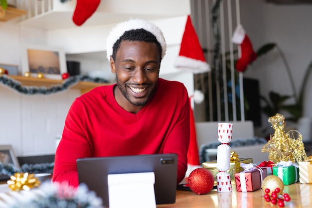 Joyeux afro-américain portant un chapeau de père Noël, utilisant une tablette pour un appel vidéo