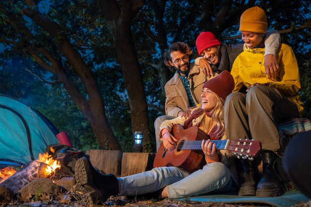 De joyeux amis multiraciaux au camping rient en feu pendant qu'une fille souriante joue de la guitare