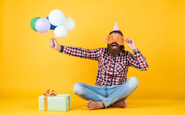 Un joyeux anniversaire détendu a l'air joyeux. homme barbu ressent la joie. homme avec boîte-cadeau célébrant l'anniversaire. hipster mature avec des ballons colorés lumineux. Fou drôle de gars.