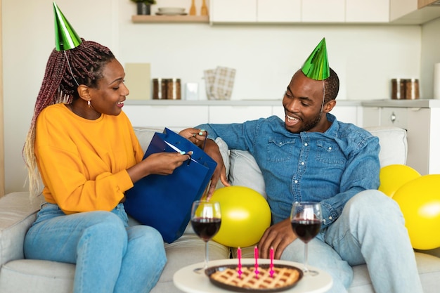 Joyeux anniversaire mon amour Femme noire excitée donnant un cadeau à son mari jeune couple assis sur un canapé à la maison