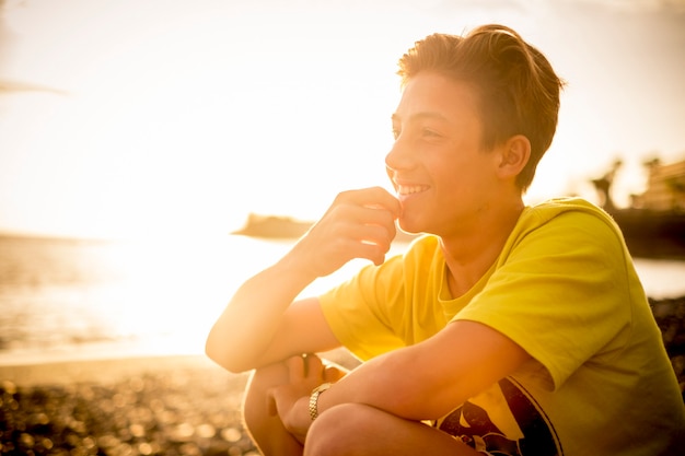 joyeux beau jeune garçon assis à la plage souriant et regardant l'horizon - des gens heureux dans l'activité de vacances de loisirs en plein air pendant le coucher du soleil doré