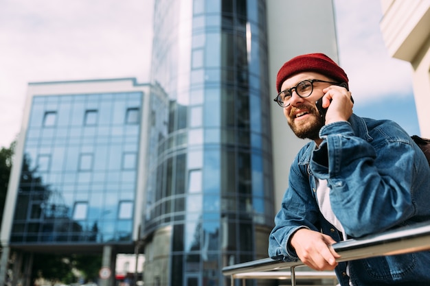 Joyeux beau jeune hipster barbu mâle parlant au téléphone et souriant. Contexte urbain avec espace copie.