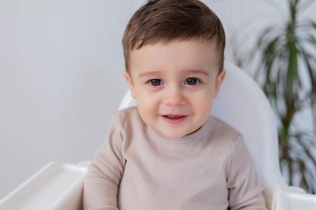 Joyeux bébé souriant est assis dans une chaise haute blanche pour nourrir fond blanc