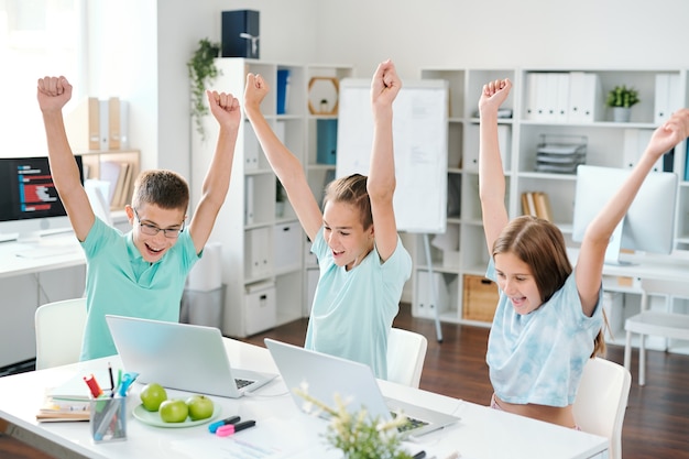 Joyeux collégiens avec des mains levées regardant des écrans d'ordinateur portable tout en exprimant leur enthousiasme