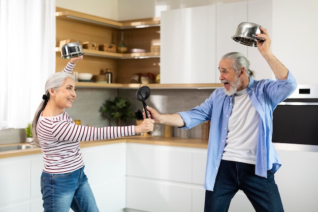 Joyeux conjoints seniors s'amusant ensemble dans la cuisine se battant de manière ludique avec des ustensiles de cuisine