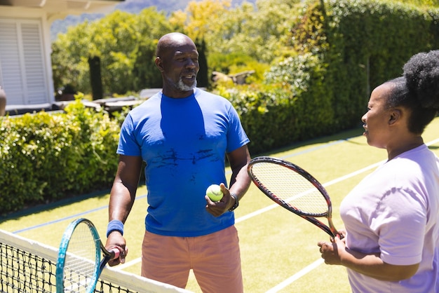 Joyeux couple afro-américain senior avec des raquettes de tennis parlant sur un terrain d'herbe ensoleillé. Mode de vie des seniors, retraite, sport, été, fitness, hobbies et loisirs.