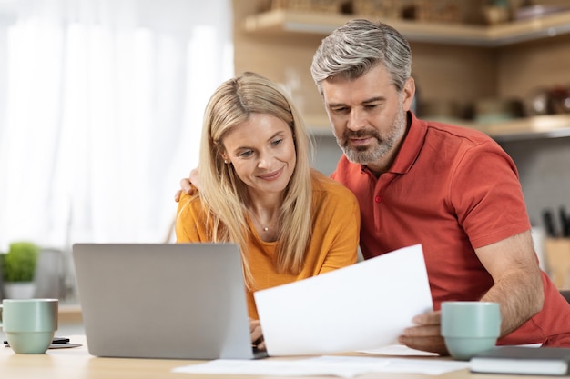 Joyeux couple d'âge moyen payant des factures en lisant des journaux