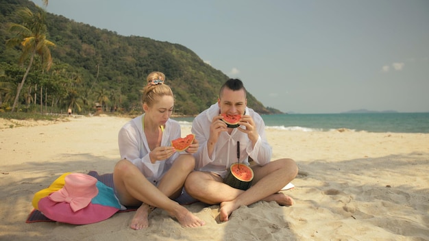 Le joyeux couple d'amoureux tenant et mangeant des tranches de pastèque sur la mer tropicale de plage de sable. Les amoureux romantiques deux personnes passent le week-end d'été.