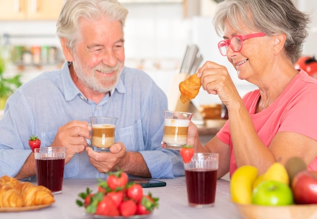 Joyeux couple caucasien prenant son petit déjeuner à la maison avec du lait, du café, un croissant. Personnes âgées détendus et heureux, appréciant la nourriture et les boissons