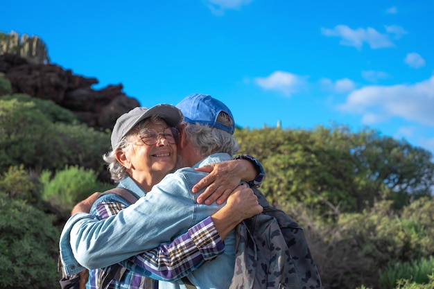 Joyeux couple caucasien senior étreignant dans la randonnée en montagne appréciant les voyages dans la nature et la liberté Sourire de vieux retraités en chapeau et vêtements décontractés parmi les buissons verts et le ciel bleu