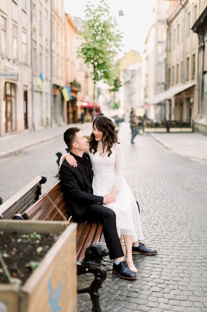 Joyeux couple de mariage chinois se tient la main tout en étant assis sur le banc dans la rue du vieux centre-ville à l'extérieur.