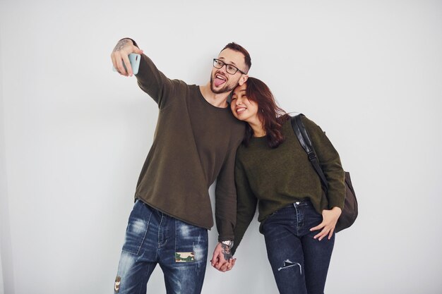 Joyeux couple multiethnique dans des vêtements décontractés faisant du selfie à l'intérieur dans le studio sur fond blanc.