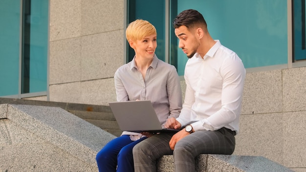 Joyeux couple multiethnique multiracial collègues arabe hispanique homme et femme caucasienne debout près du centre d'affaires dans la rue à l'extérieur et travaillant sur un ordinateur portable discuter du projet analyser le travail d'équipe de données