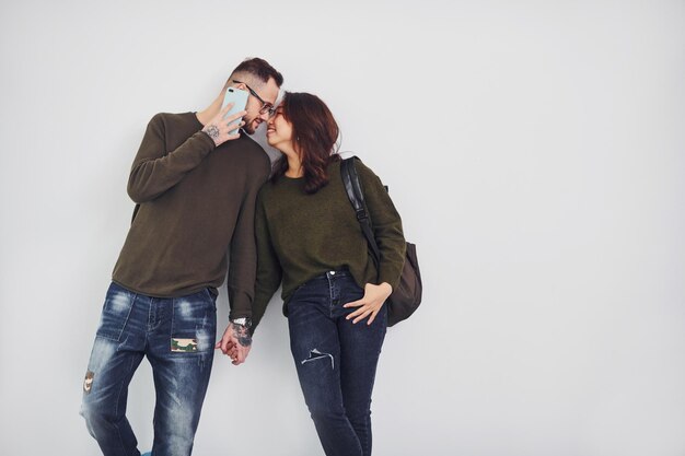 Joyeux couple multiethnique avec sac à dos et téléphone debout ensemble à l'intérieur dans le studio sur fond blanc.