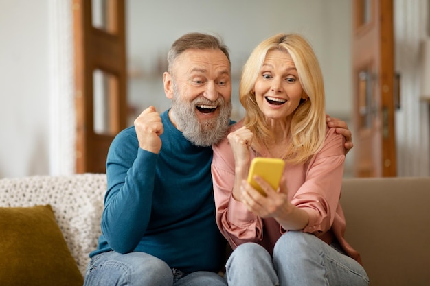 Joyeux couple de personnes âgées à l'aide d'un message de lecture de smartphone secouant les poings à l'intérieur