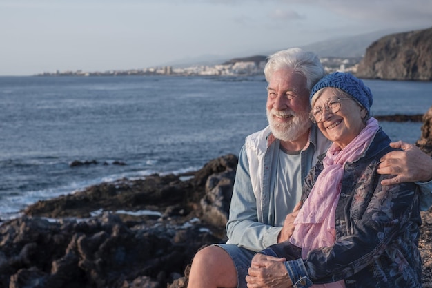 Joyeux couple de personnes âgées charmant assis sur les rochers en mer en profitant de la lumière du coucher du soleil