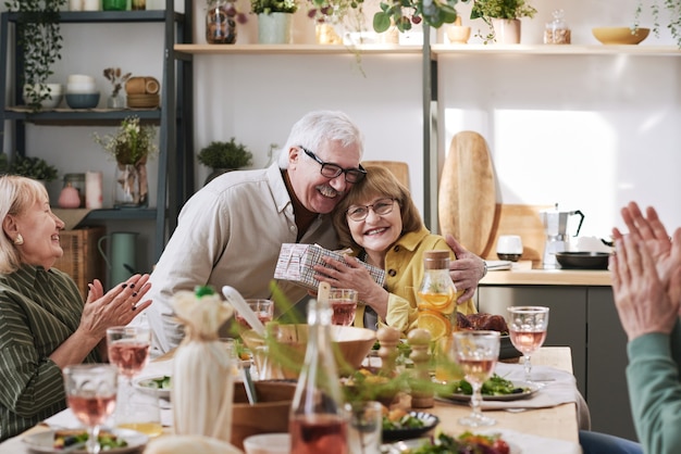 Joyeux couple de personnes âgées tenant un cadeau et s'embrassant tout en célébrant avec des amis à la table à manger