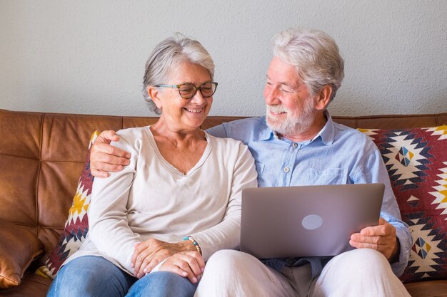 Joyeux couple de personnes âgées utilisant un ordinateur portable assis sur un canapé et souriant. Couple de personnes âgées heureux se détendre en surfant sur un ordinateur portable assis dans le salon. Vieux couple regardant du contenu multimédia à l'aide d'un ordinateur portable