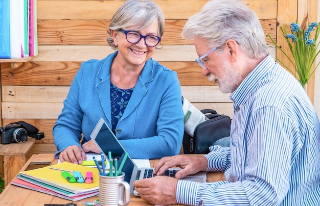 Joyeux couple de personnes aux cheveux blancs assis en plein air à une table en bois travaillant avec un ordinateur portable.