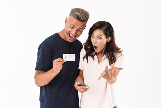 Joyeux couple séduisant portant une tenue décontractée debout isolé sur un mur blanc, faisant des achats en ligne avec un téléphone portable et une carte de crédit