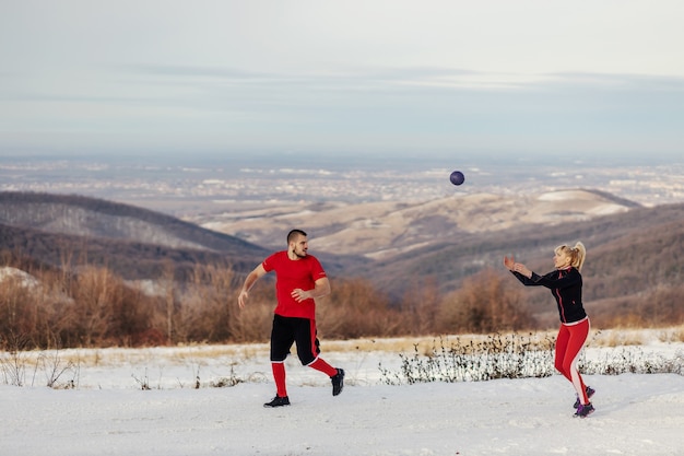 Joyeux couple sportif faisant du jogging dans la nature et jetant vers avec un ballon de fitness le jour d'hiver enneigé. Fitness d'hiver, vie saine, fitness en plein air
