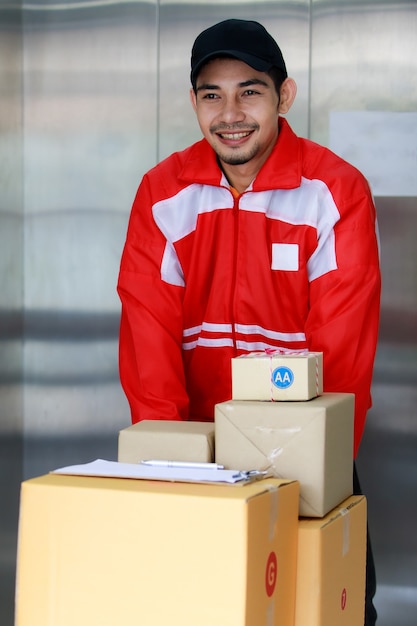 Un joyeux courrier asiatique pousse une pile de boîtes en carton sur un chariot hors de l'ascenseur. Livraison de colis dans un immeuble résidentiel moderne en zone urbaine. Concept d'entreprise de commerce électronique.