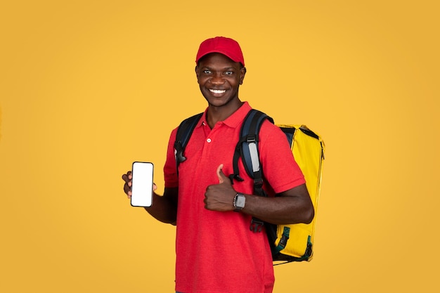 Joyeux coursier afro-américain millénaire avec sac à dos et téléphone avec écran vide pouce vers le haut