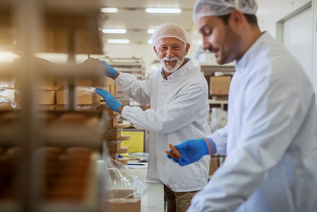 Joyeux employés caucasiens vêtus d'uniformes stériles blancs emballant des biscuits dans des boîtes en se tenant debout dans une usine alimentaire.