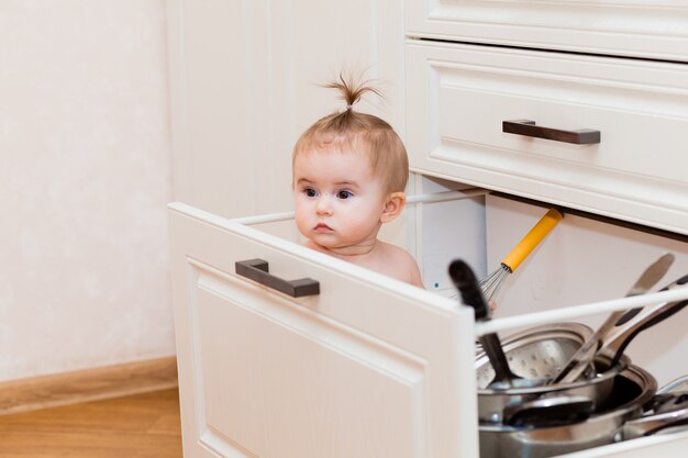 Joyeux enfant assis dans le tiroir de la cuisine avec des casseroles et riant. Portrait d'un enfant dans une cuisine blanche.